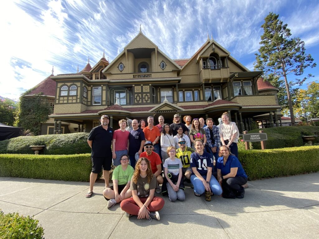 A group of people poses in front of the Winchester Mystery House, a large Victorian-style mansion with ornate architecture and a beautifully landscaped garden.