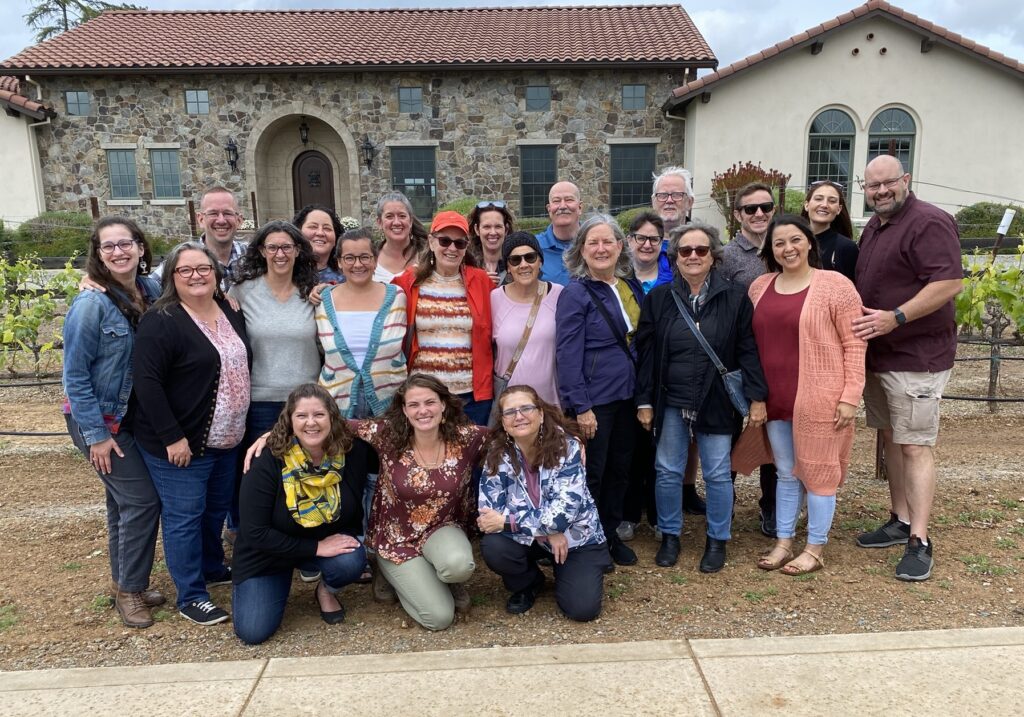 A large group of smiling people poses in front of a rustic stone winery building with a red-tiled roof, surrounded by grapevines.