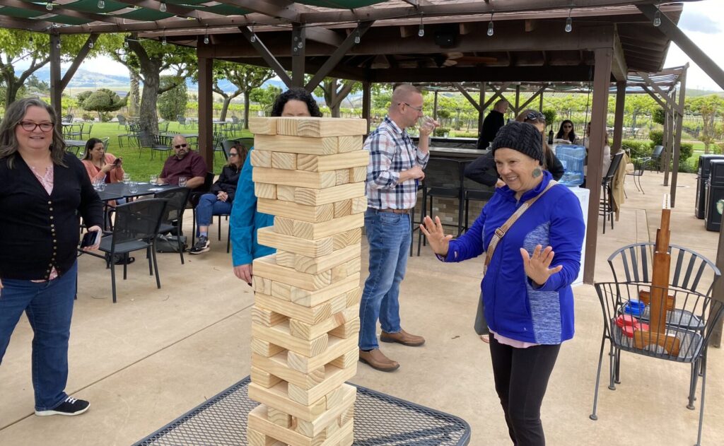 A woman in a blue jacket stands near a giant Jenga tower, smiling and raising her hands as if about to make a move. Others watch in the background at an outdoor winery setting.
