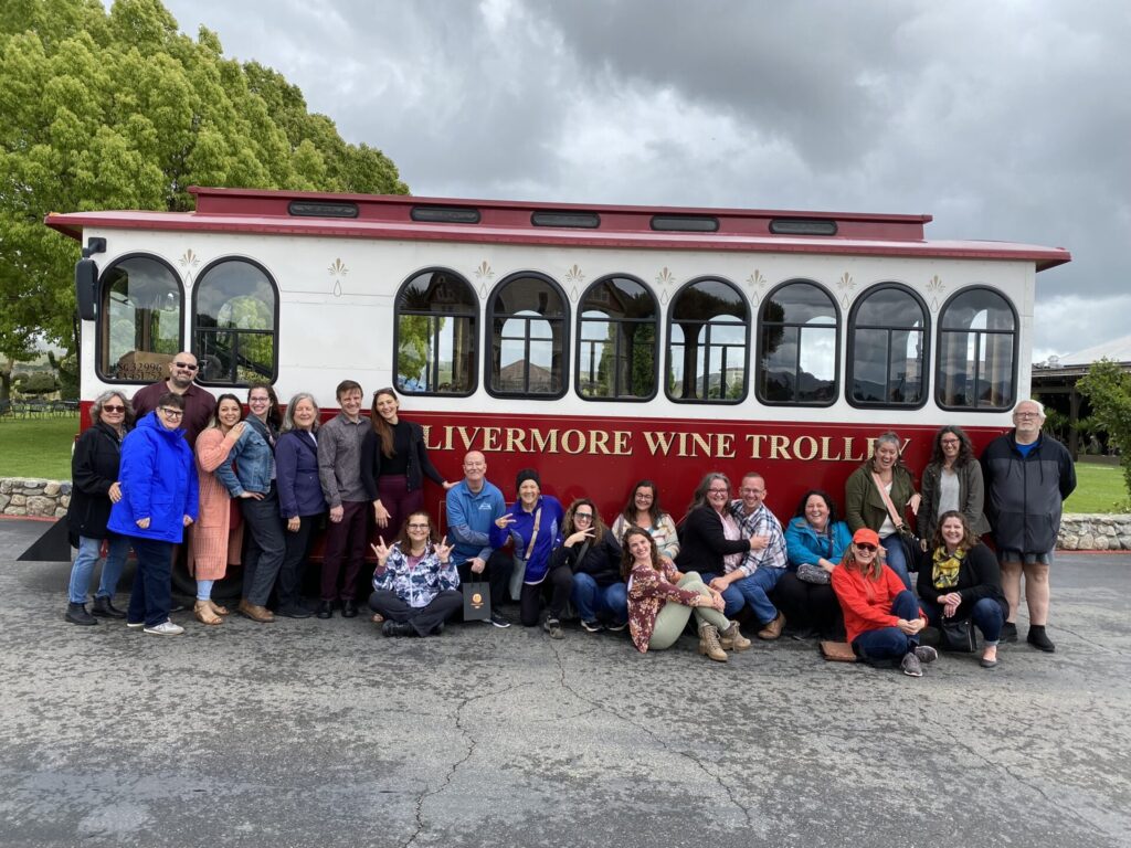 A large group of people poses in front of the “Livermore Wine Trolley,” a red and white vintage-style trolley. The group is diverse, with some sitting, kneeling, or standing, making various hand gestures and smiles.