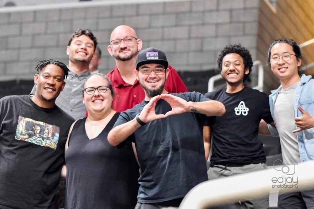 A group of seven people poses together in an indoor stadium setting, smiling at the camera. One person in the center forms a heart shape with their hands. The image has a watermark reading “Ed Jay Photography.”