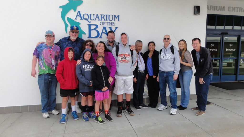 A diverse group of adults and children poses outside the entrance of “Aquarium of the Bay.” The sign with a blue shark logo is visible above them.