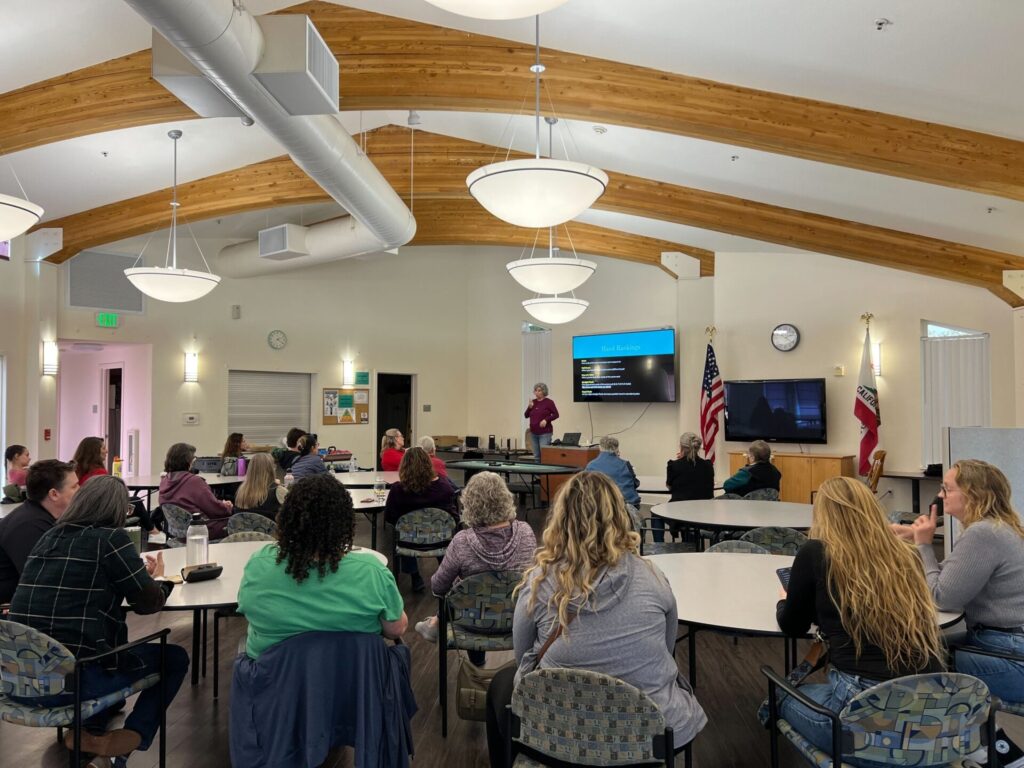 A speaker in a purple shirt presents to a seated audience in a well-lit community room with wooden beams and American and California flags. A large screen displays a slide.
