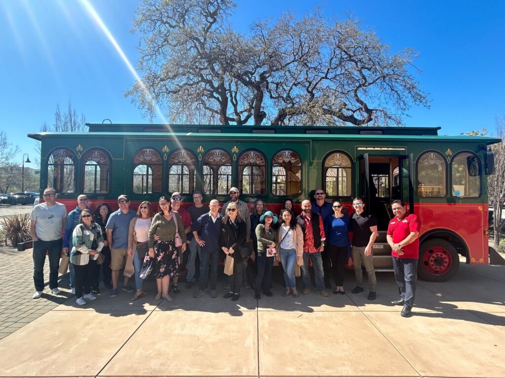 A large group poses in front of a vintage-style green and red trolley under a bright blue sky, with trees in the background.