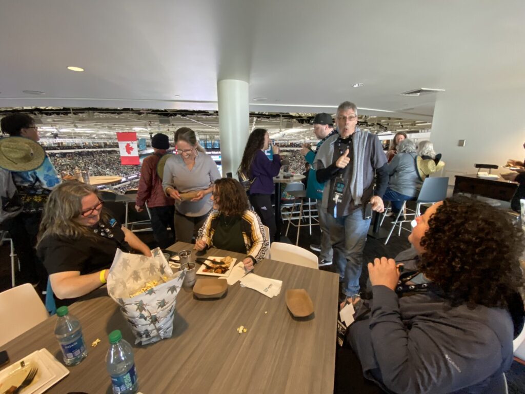 A group of people enjoys food and conversation in a VIP lounge overlooking a sports arena, with a Canadian flag visible in the background.