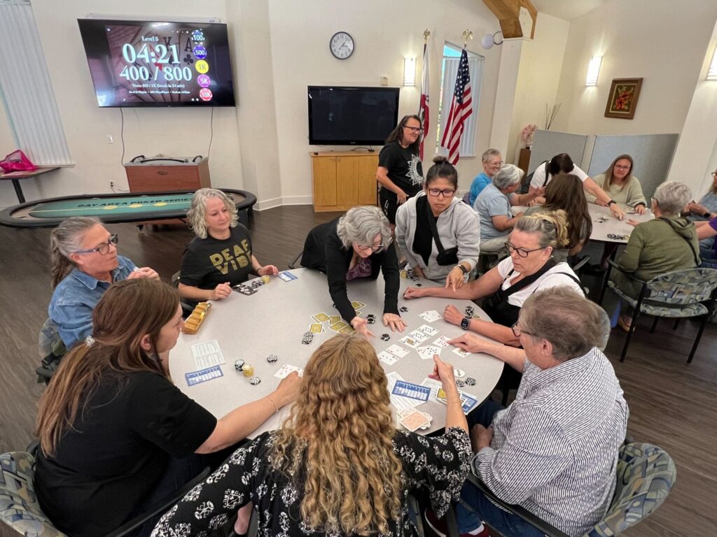 A group of people is engaged in a poker game at a community event, with playing cards and poker chips spread across tables. A timer and betting amounts are displayed on a screen in the background.