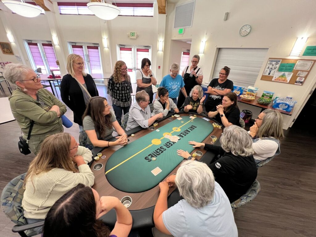 A group of women sits around a poker table with chips and cards, while others stand behind them, watching the game.