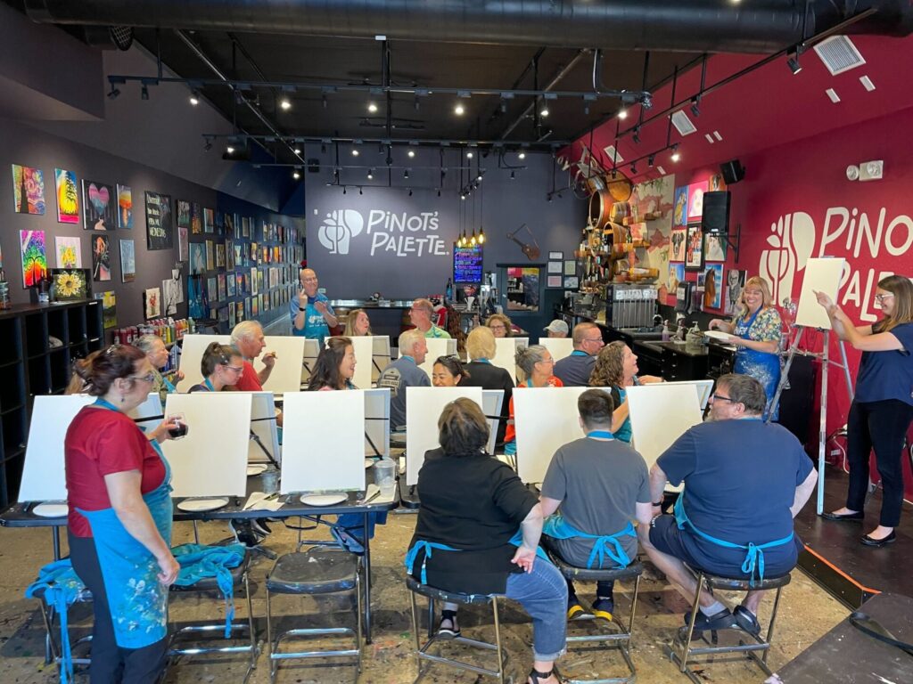 A large group participates in a painting event at Pinot’s Palette, seated in front of blank canvases. The venue has colorful artwork on the walls and instructors at the front.