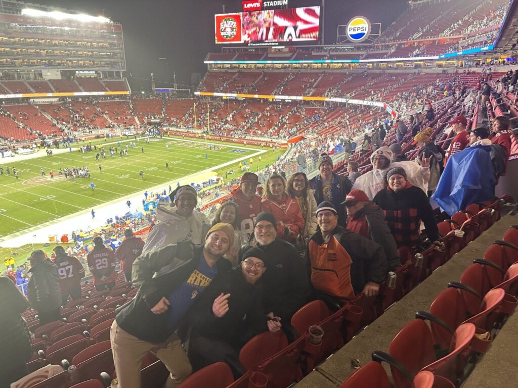 A group of football fans poses in a stadium with a game playing on the field below. They are dressed in warm clothing, some in team colors.