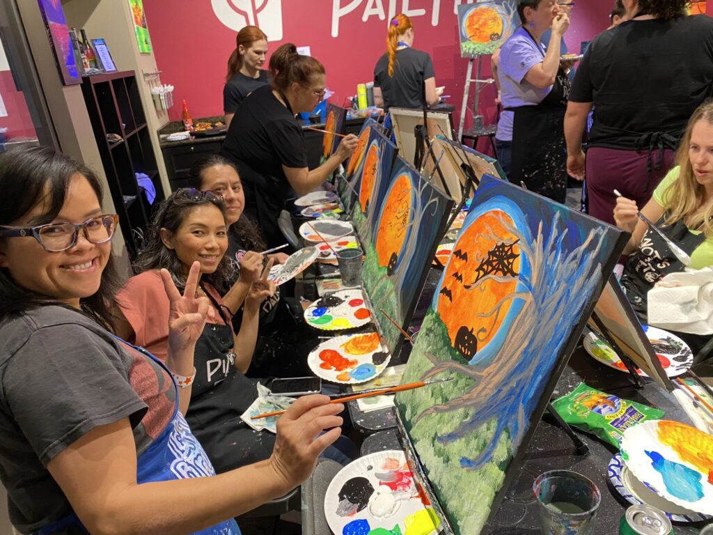 A group of women at a painting event, smiling and holding brushes while working on colorful Halloween-themed paintings. The setting has a pink wall with “PAINT” in white letters.