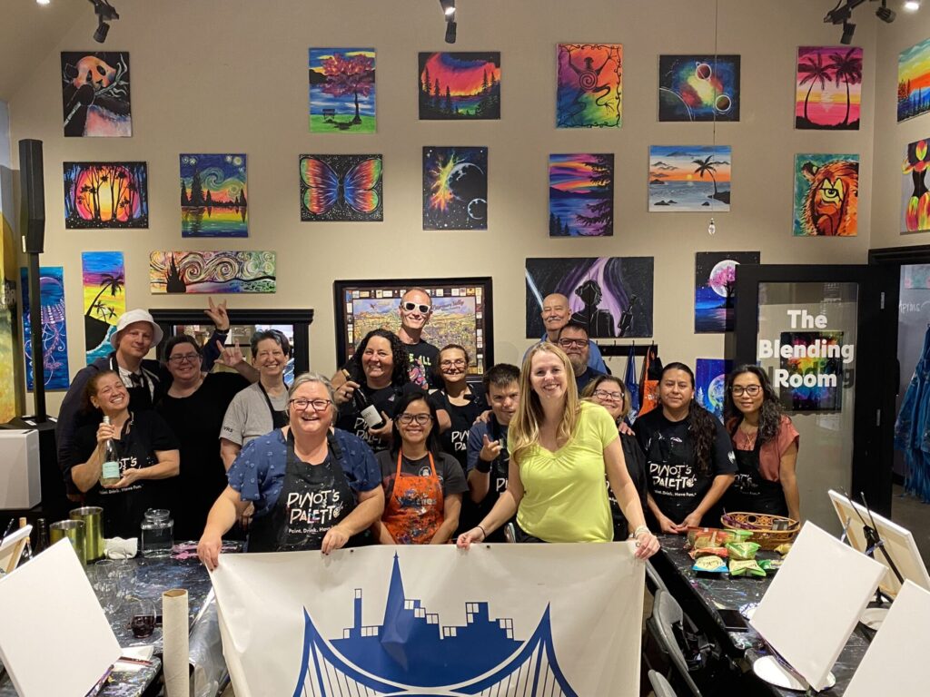 A group of people at a painting studio posing together in front of colorful artwork. They hold a banner with a blue bridge logo and smile. The walls display vibrant paintings, including a butterfly, landscapes, and abstract art.