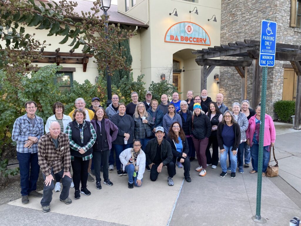 A large group of people pose outside a venue called “Da Boccery,” standing near greenery and a wooden pergola.