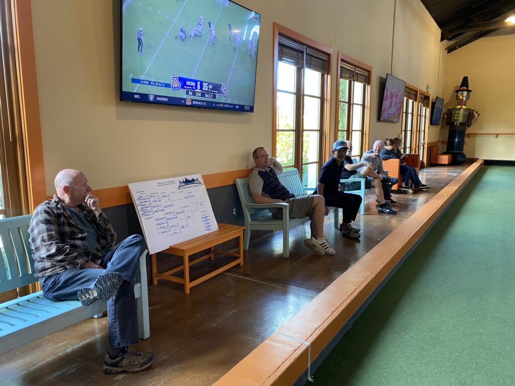 Several men sit on benches watching an indoor bocce game. A tournament bracket is displayed on a board nearby, and a large TV screen shows a football game.