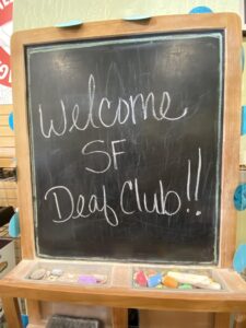 A chalkboard sign with the handwritten message “Welcome SF Deaf Club!!” displayed in a wooden frame with colorful chalk pieces at the bottom.