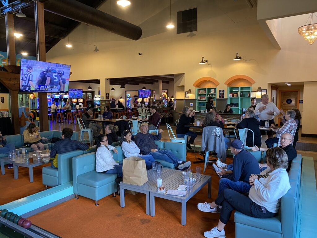 A spacious venue with people socializing, sitting on blue couches, and playing arcade games. A football game is playing on a large TV screen.