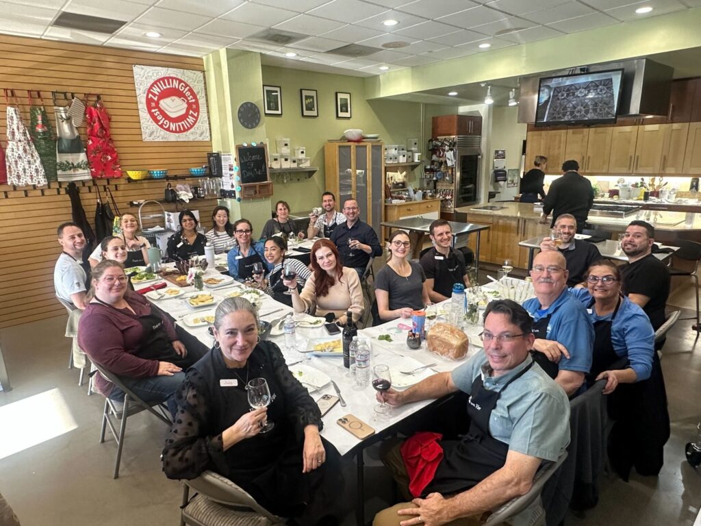 A large group of people sit around a long table in a kitchen setting, smiling and holding drinks. A sign in the background reads “Zwilling.”