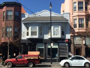 A modern daytime photo of a two-story building with bay windows and a gray facade, with a business called “HOUSE” on the ground floor.