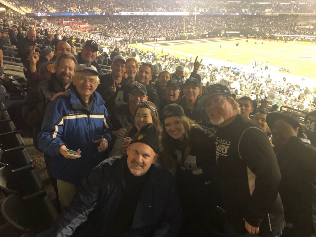 A group of football fans poses in a stadium with a game playing on the field below. They are dressed in warm clothing, some in team colors.