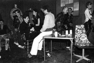A black-and-white photo of a punk club scene with people sitting and standing around a table with drinks. Some wear leather jackets and punk attire.