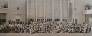 A sepia-toned vintage photograph of a large group of people gathered in front of a modernist-style building with tall windows and decorative light fixtures. Many are dressed in formal attire, with hats and suits. A small sign in the center reads “National Association of the Deaf.”