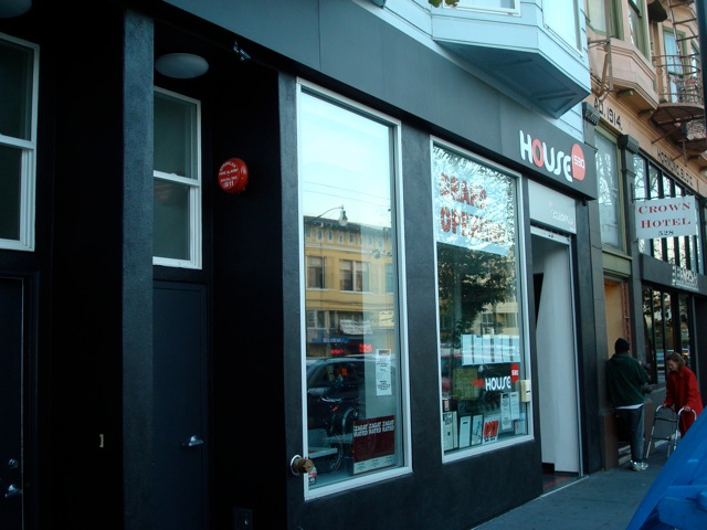 A close-up view of the entrance to “HOUSE,” a modern storefront with large glass windows framed in black. The interior is visible through the windows.